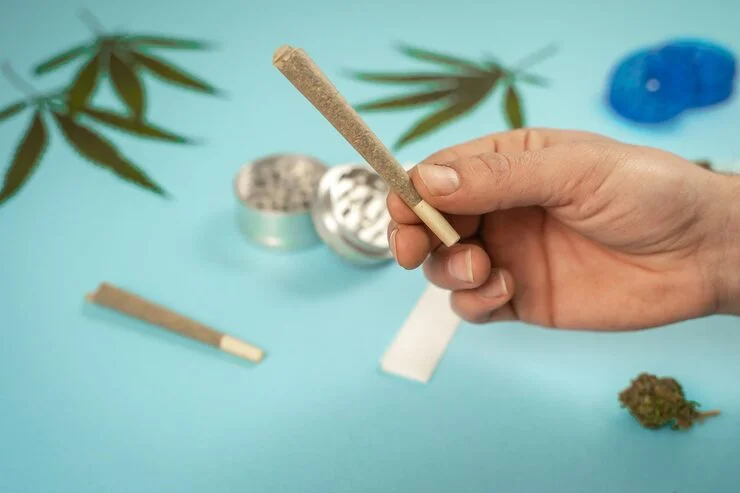 Hand holding a rolled cannabis joint with marijuana leaves, grinder, and accessories on a turquoise background.