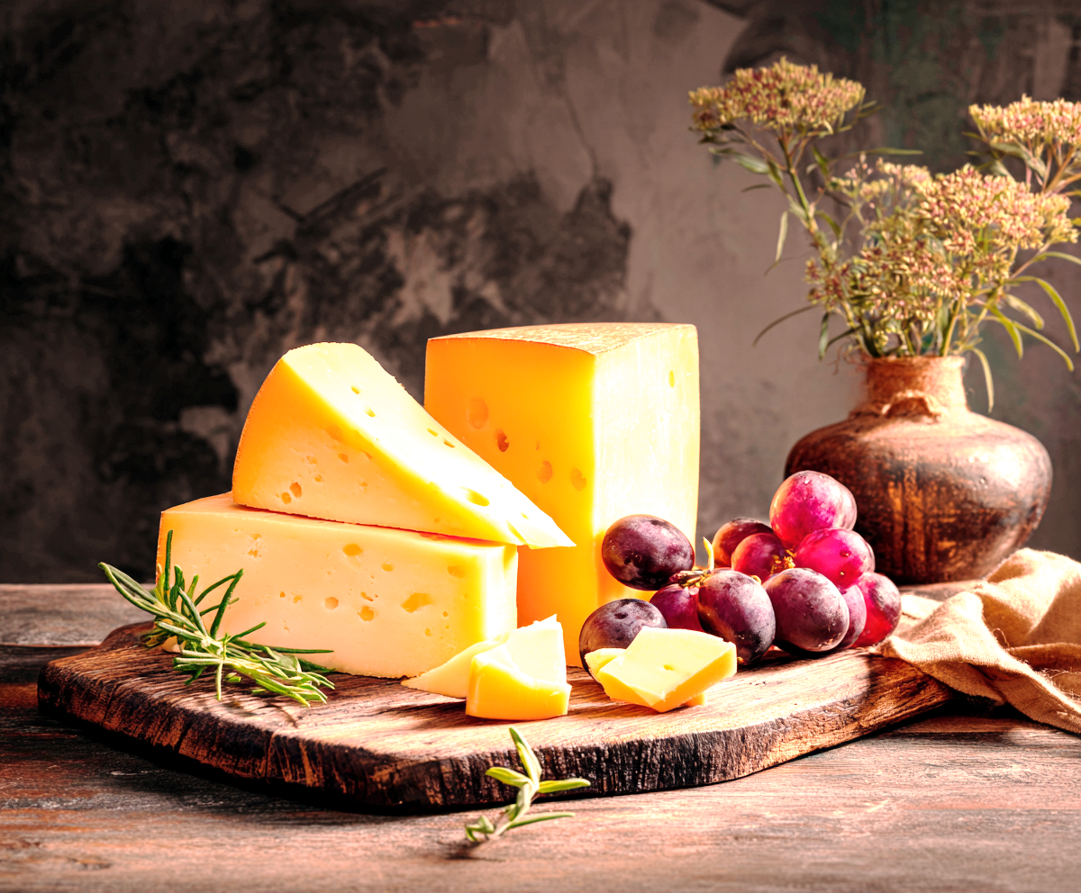 A rustic still life arrangement of various cheese wedges on a wooden board with red grapes and fresh rosemary, set against a dark textured background with a ceramic vase of flowers.