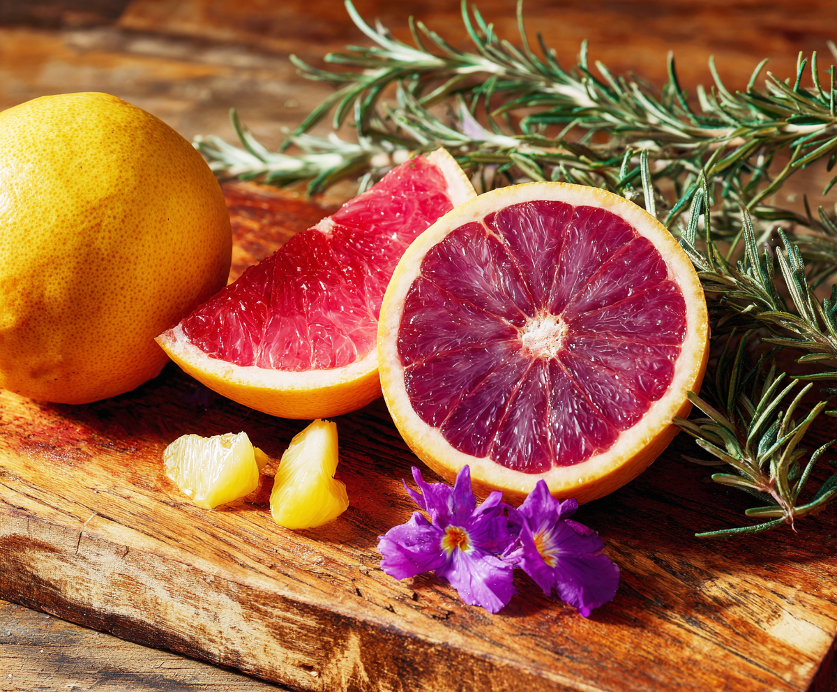 Sliced ruby grapefruit and rosemary sprigs on a rustic wooden board with purple flowers.