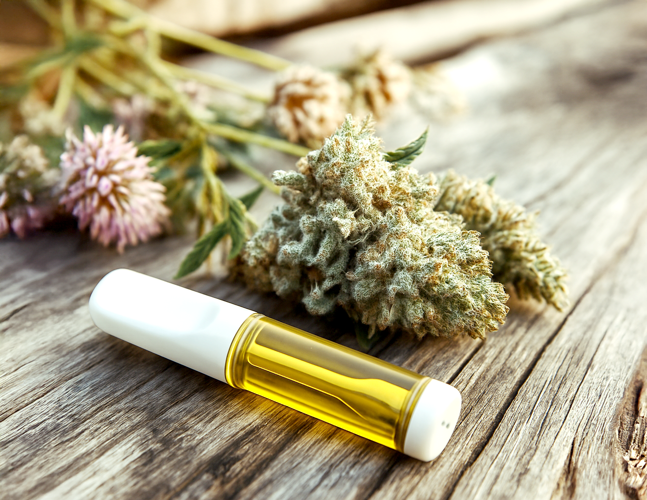 A cannabis vape cartridge placed beside dried cannabis buds and blooming clover flowers on a rustic wooden surface.