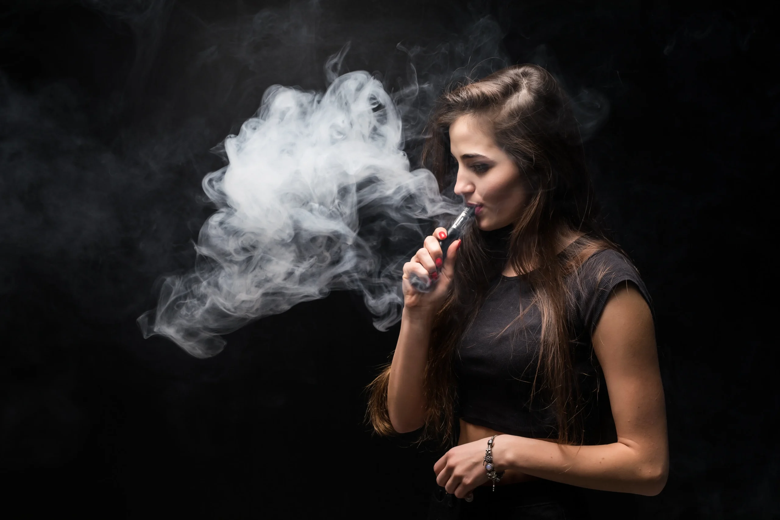 A young woman in a black top exhales a thick cloud of vapor while using a vape pen against a dark background.