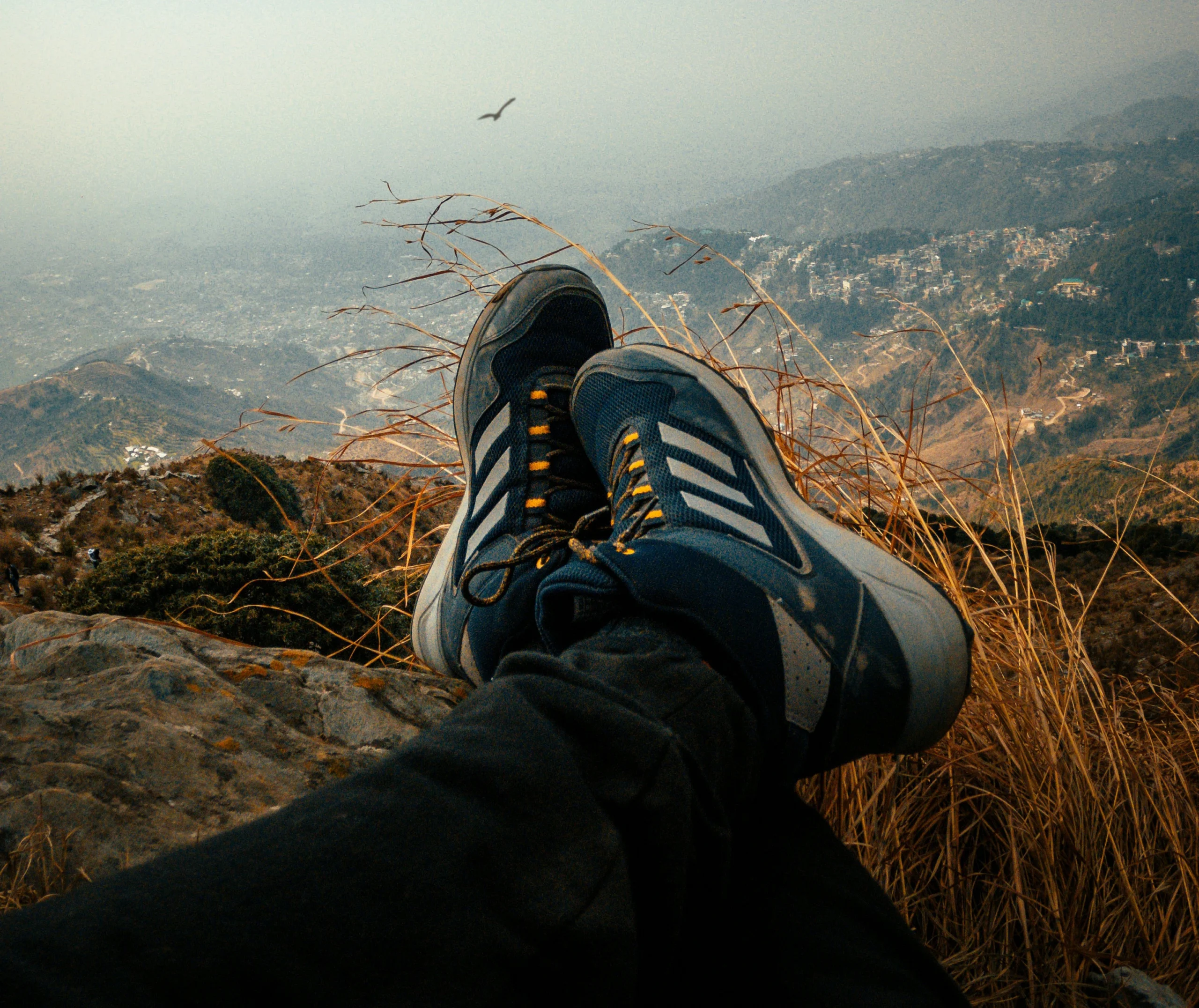 Person’s legs in dark pants with blue hiking shoes resting on a rocky cliff overlooking a mountainous valley.